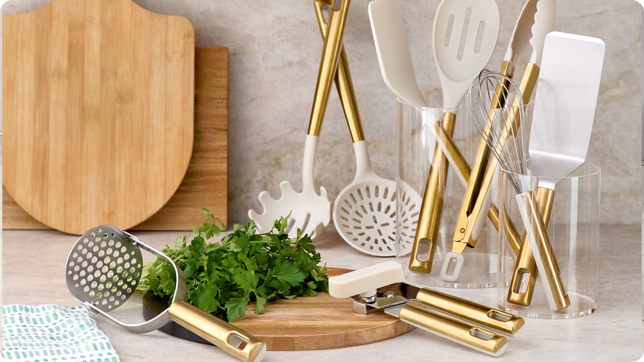 Simmer & Stir campaign kitchen styling featuring gold‑and‑white cooking utensils, silicone spatulas, whisk, slotted spoon, and clear utensil holders arranged with wooden cutting boards and fresh herbs on a countertop.